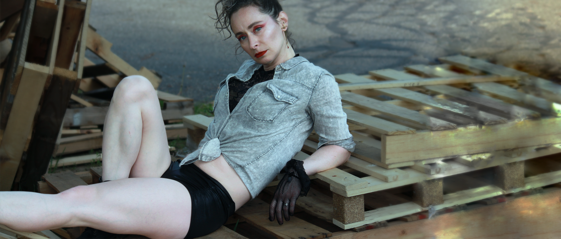 Woman in shiny black metallic pole shorts reclines on wooden pallets